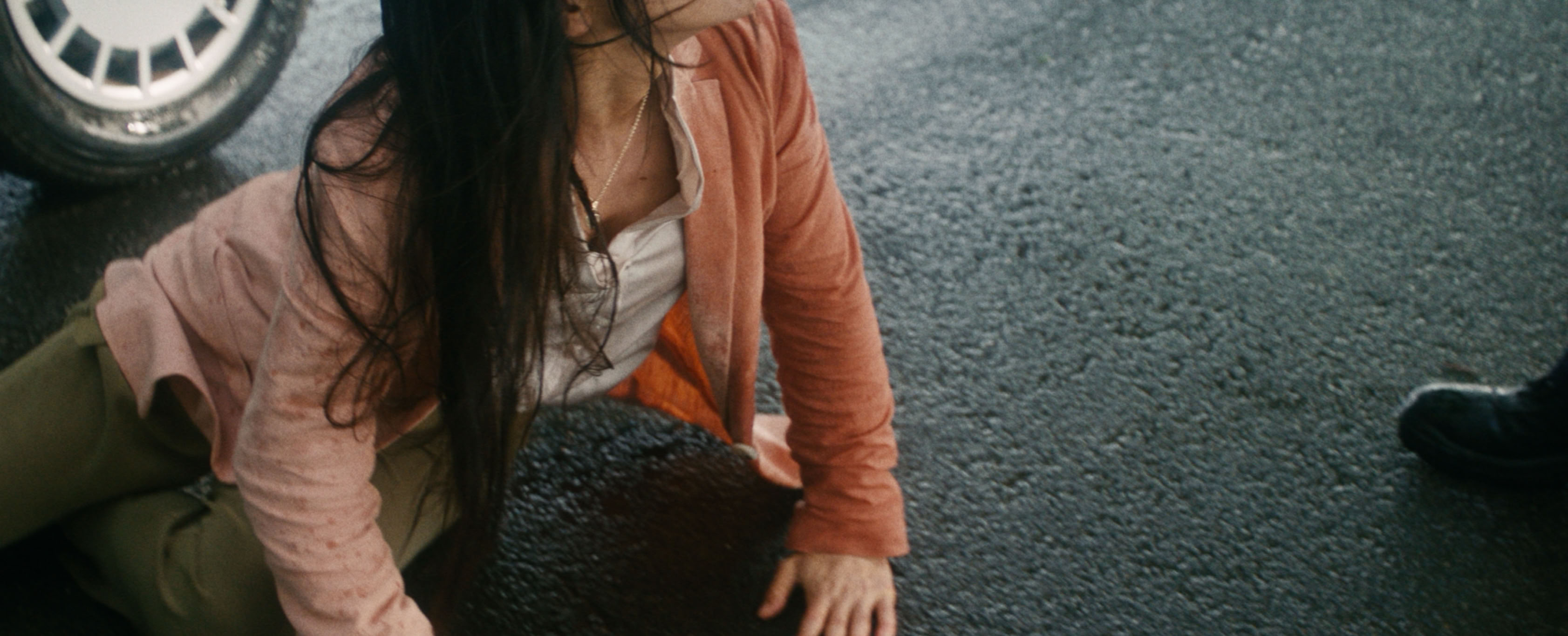 A person with long hair wearing a pink jacket and white shirt is on their hands and knees on a wet road surface, near a vehicle tire.